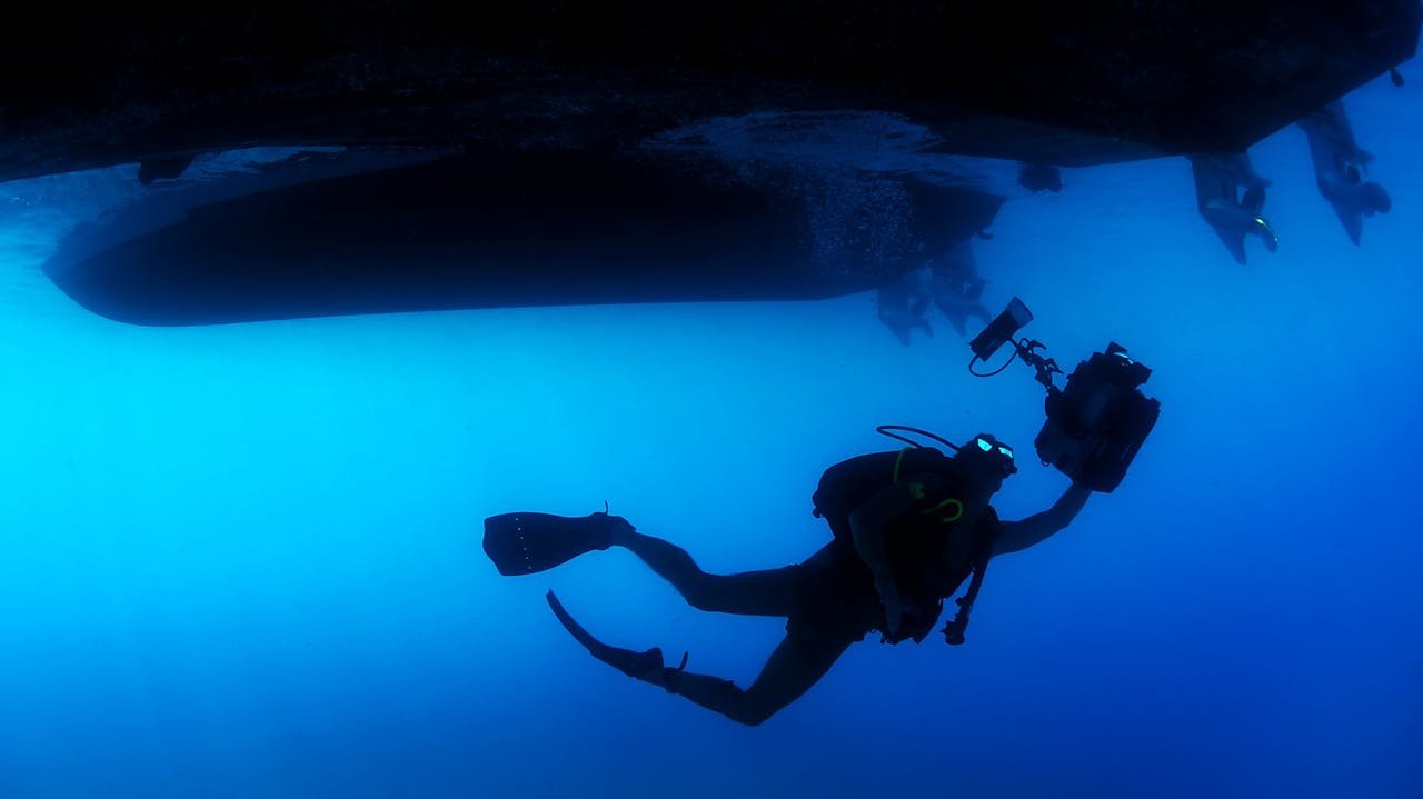 A diver with a camera explores the ocean depths beneath a boat.